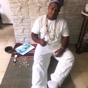 Puede incluir: Una persona con una camiseta blanca, pantalones blancos y un tocado decorativo morado y dorado. La persona está sentada en una alfombra marrón y verde, con una pintura, conchas y un cuenco pequeño cerca.