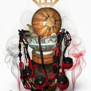 Puede incluir: Una calabaza redonda descansa en un cuenco de cerámica estampado sobre un pedestal de madera. Cuentas negras y rojas con pequeñas esferas negras y conchas de cauri cuelgan alrededor de la base. Humo rojo y gris rodea el objeto con un icono dorado.