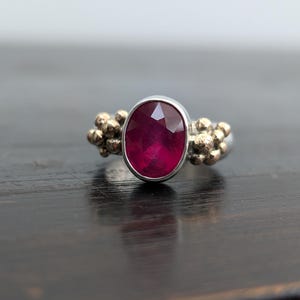 May include: A silver ring with a large, oval-cut ruby gemstone. The ruby is a deep red color and is set in a silver bezel. The ring has gold-colored accents on either side of the ruby. The ring is displayed on a dark wooden surface.