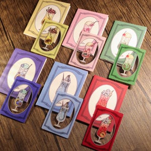 Puede incluir: Colección de cuadernos y tarjetas coloridas con ilustraciones de diversas bebidas en capas. Los cuadernos y tarjetas son en tonos amarillo, rosa, verde, morado, azul y rojo, sobre un fondo de madera.