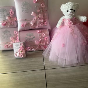 May include: A collection of pink decorative items, including a pillow, boxes, and a teddy bear. The items are adorned with floral accents, bows, and text. The teddy bear wears a pink dress and a crown.