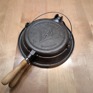 May include: A round cast iron waffle iron with two light-coloured wooden handles and a wire bail handle. The top surface features the embossed text Taylor Forbes Co Ltd, Guelph, Canada, and a diamond-shaped logo with the letters TF inside.