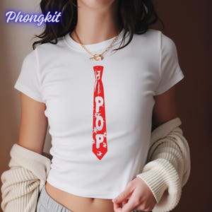 Può includere: Top corto bianco con grafica di cravatta rossa con la scritta "POP" in lettere bianche. Il design della cravatta include accenti a forma di stella. La maglietta è a maniche corte e ha un girocollo.