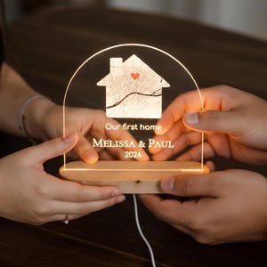 May include: A personalized LED night light with a house-shaped design and a map graphic. The text "Our first home" is above the names "MELISSA & PAUL" and the year "2026". The light has a wooden base and a clear acrylic top.