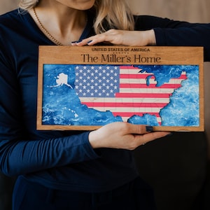 May include: Wooden framed artwork featuring a map of the United States with the American flag design. The map includes the states, Alaska, and Hawaii, set against a blue background. The text "UNITED STATES OF AMERICA" and "The Miller's Home" are displayed.