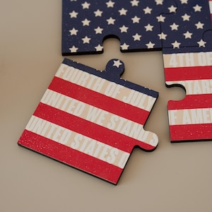 Peut inclure: Pièces de puzzle en bois sur le thème patriotique. Une pièce affiche des rayures rouges et blanches avec le texte "United States" et "Fourth of July". D'autres pièces montrent un fond bleu avec des étoiles blanches.