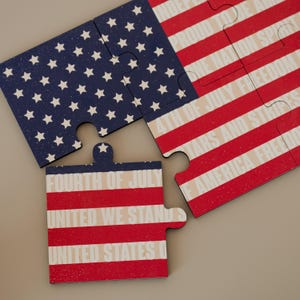 Peut inclure: Puzzle de quatre pièces représentant le drapeau américain. Les pièces montrent les étoiles et les rayures du drapeau, avec des phrases patriotiques comme "Fourth of July" et "United We Stand". Les couleurs sont le rouge, le blanc et le bleu.