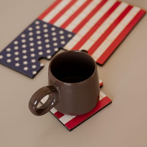 Peut inclure: Une tasse en céramique marron avec une anse incurvée repose sur un dessous de verre à rayures rouges et blanches. Un puzzle en bois du drapeau américain est partiellement assemblé, avec des étoiles bleues et des rayures rouges.