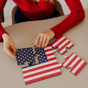 Peut inclure: Un puzzle en bois représentant le drapeau américain, avec des rayures rouges et blanches et un champ bleu d'étoiles blanches. Le puzzle est partiellement assemblé, avec des pièces en cours de connexion. Le texte "UNITED STATES OF AMERICA" est visible.