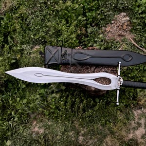 Puede incluir: Una espada plateada y negra con un mango y guarda decorativos, apoyada sobre una superficie de madera con una vaina negra. La hoja tiene un diseño recortado único. La espada se exhibe sobre un lecho de follaje verde.