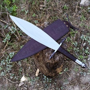 May include: A handmade sword with a silver blade and a dark brown leather-wrapped handle. The sword rests on a wooden stump, with a brown leather sheath. The sword's design features a twisted guard and a silver pommel.