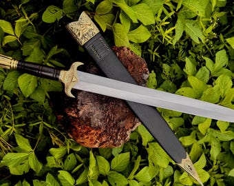 May include: A decorative sword with a silver blade and gold accents, resting on a wooden surface. The sword's handle is black and gold, and it is accompanied by a black sheath. The background is a bed of green leaves.