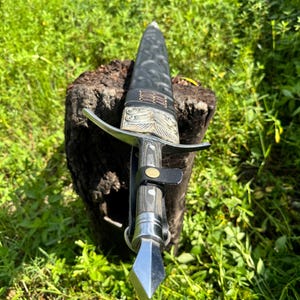 May include: A dagger with a silver-toned metal pommel and crossguard, a dark wood-grain handle, and a black leather sheath. The sheath features intricate embossed patterns, a silver-toned metal throat, and a black leather strap with a brass snap.