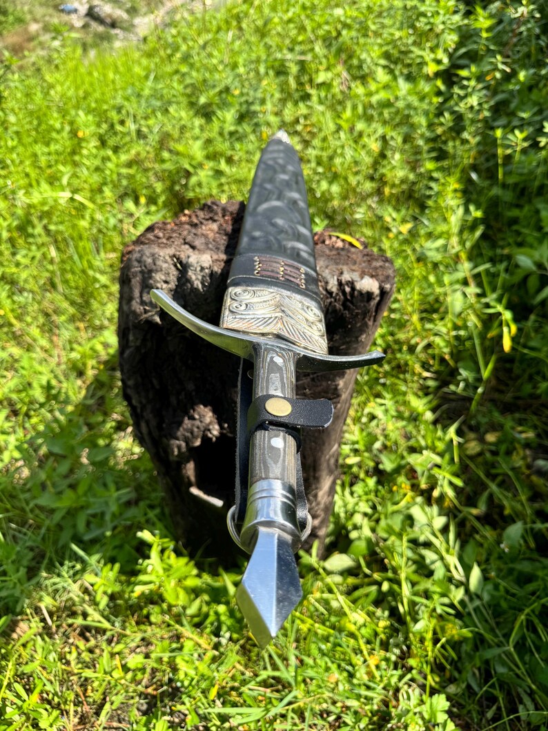 May include: A dagger with a silver-toned metal pommel and crossguard, a dark wood-grain handle, and a black leather sheath. The sheath features intricate embossed patterns, a silver-toned metal throat, and a black leather strap with a brass snap.