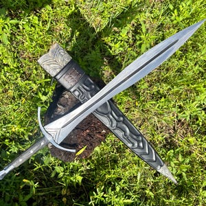 May include: A double-edged steel sword with a pointed tip, a central fuller, and a crossguard. The handle is dark with metallic accents. It is positioned over a black scabbard featuring intricate embossed patterns and a metallic top fitting.