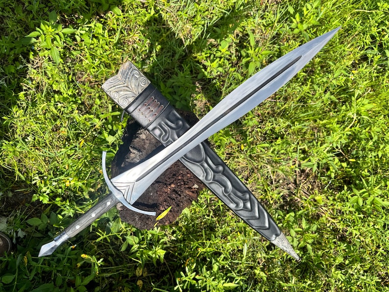 May include: A double-edged steel sword with a pointed tip, a central fuller, and a crossguard. The handle is dark with metallic accents. It is positioned over a black scabbard featuring intricate embossed patterns and a metallic top fitting.