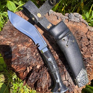 May include: A large, curved knife with a polished steel blade and a dark wooden handle. The knife is accompanied by a black leather sheath with a belt loop and decorative metal accents. The knife and sheath are displayed on a piece of wood.
