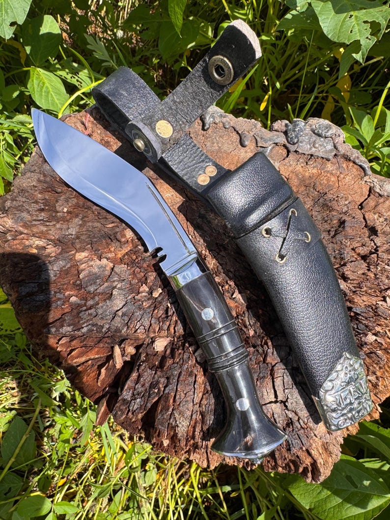 May include: A large, curved knife with a polished steel blade and a dark wooden handle. The knife is accompanied by a black leather sheath with a belt loop and decorative metal accents. The knife and sheath are displayed on a piece of wood.