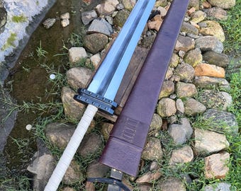 May include: A large, silver-bladed sword with a white wrapped handle and black accents. The sword is paired with a brown leather sheath and a black leather hilt. The sword is lying on a bed of rocks and grass.