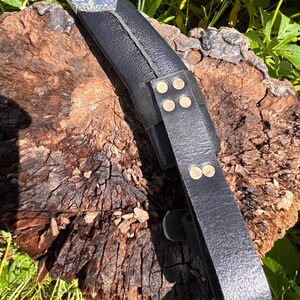 May include: A black leather sheath with a silver and decorative top. The sheath has a strap with brass rivets. The object is resting on a weathered, brown wooden surface, with green foliage in the background.