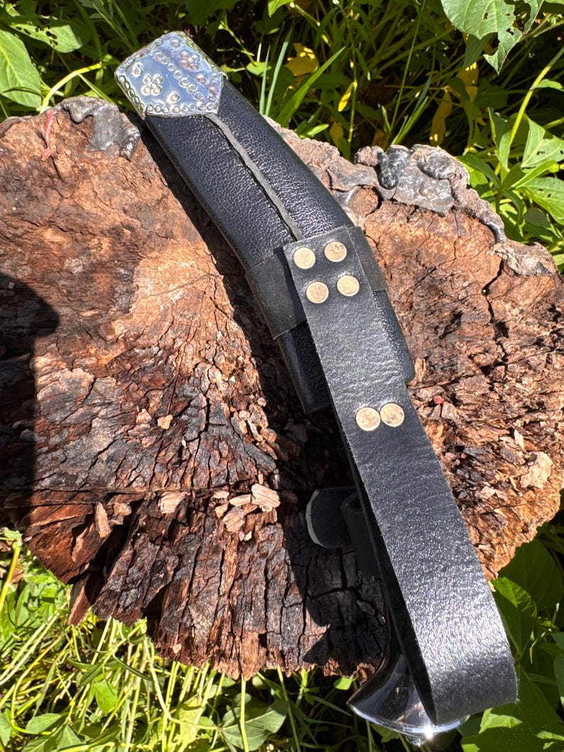 May include: A black leather sheath with a silver and decorative top. The sheath has a strap with brass rivets. The object is resting on a weathered, brown wooden surface, with green foliage in the background.