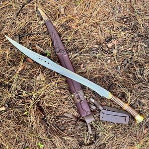 May include: A double-edged sword with a curved, silver-toned blade featuring four circular cutouts near the hilt. The handle is ribbed with a gold-coloured pommel. It is accompanied by a brown leather scabbard with gold-coloured metal accents.