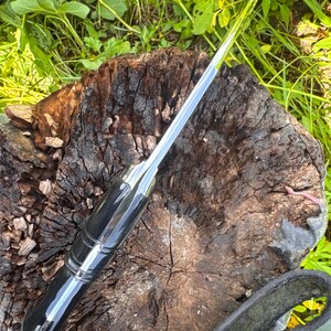 May include: A polished, silver-bladed knife with a black handle. The knife is resting on a weathered, brown tree stump, with green foliage in the background. The knife has a sleek, modern design.