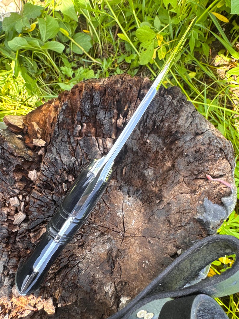 May include: A polished, silver-bladed knife with a black handle. The knife is resting on a weathered, brown tree stump, with green foliage in the background. The knife has a sleek, modern design.