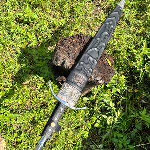 May include: A dagger with a black sheath featuring embossed patterns and a silver-colored metal throat. The handle has a silver crossguard, a dark grip with a central gold-colored rivet, and a silver pommel. The sheath has a leather-like texture.