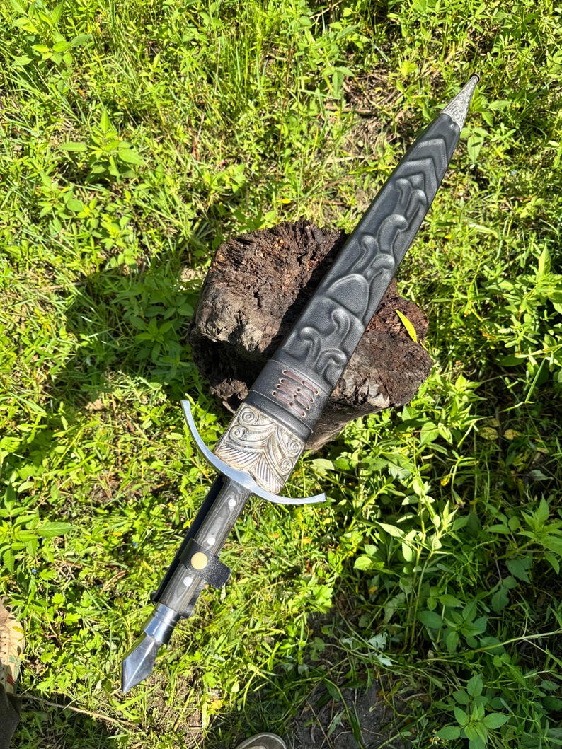May include: A dagger with a black sheath featuring embossed patterns and a silver-colored metal throat. The handle has a silver crossguard, a dark grip with a central gold-colored rivet, and a silver pommel. The sheath has a leather-like texture.
