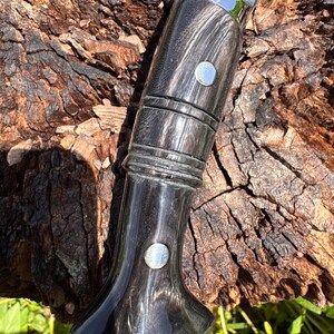 May include: Close-up of a knife handle with a dark, wood-grain finish. The handle has a curved shape with two silver rivets and a polished, reflective surface. The blade is silver and the knife is resting on a textured, brown surface.