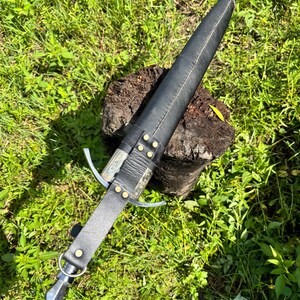 May include: A dagger encased in a black leather sheath with brass rivets and a metal crossguard. The handle is wrapped in black leather and features a metal pommel with a ring attachment. The sheath has a pointed metal tip at the end.