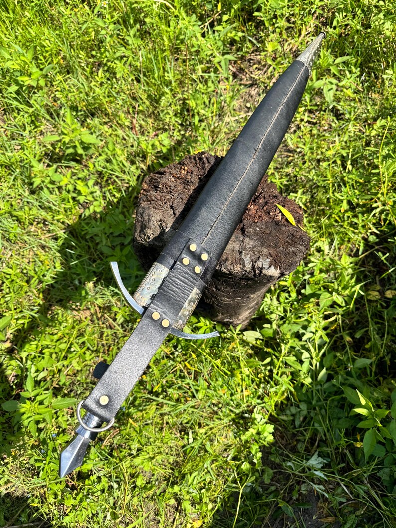 May include: A dagger encased in a black leather sheath with brass rivets and a metal crossguard. The handle is wrapped in black leather and features a metal pommel with a ring attachment. The sheath has a pointed metal tip at the end.