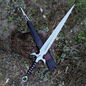 May include: A silver and black sword with a pointed blade and a decorative hilt, resting on a weathered wooden surface. The sword is sheathed in a black scabbard with red accents. The overall design suggests a fantasy or historical theme.