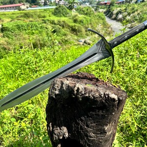 May include: A double-edged steel sword featuring a pointed blade with a central fuller, a curved metal crossguard with decorative V-shaped detailing, and a dark wooden handle secured with metal rivets and a pointed pommel at the end.