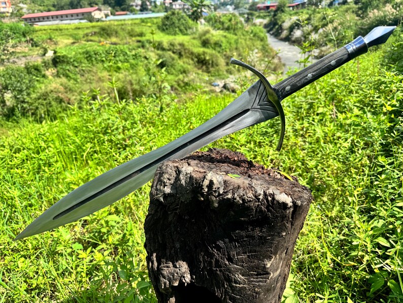 May include: A double-edged steel sword featuring a pointed blade with a central fuller, a curved metal crossguard with decorative V-shaped detailing, and a dark wooden handle secured with metal rivets and a pointed pommel at the end.