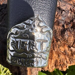 May include: Close-up of a silver-colored metal object with engraved text and floral designs. The object is attached to a black leather surface. The text appears to be in a foreign language.