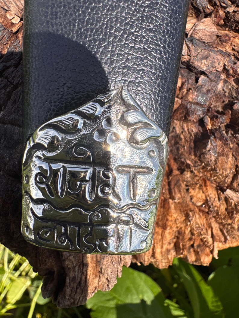 May include: Close-up of a silver-colored metal object with engraved text and floral designs. The object is attached to a black leather surface. The text appears to be in a foreign language.