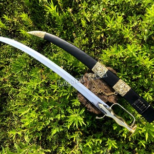 May include: A curved sword with a polished silver blade and a metallic hilt, accompanied by a black scabbard featuring ornate gold-coloured metal fittings with intricate scrollwork patterns and a small hanging chain attached to the side.