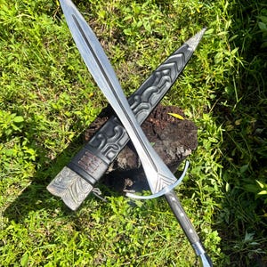 May include: A double-edged steel sword with a central fuller and a silver-toned crossguard. The handle is dark wood with metal rivets. It is accompanied by a black leather scabbard featuring embossed scrollwork, metal fittings, and leather lacing.