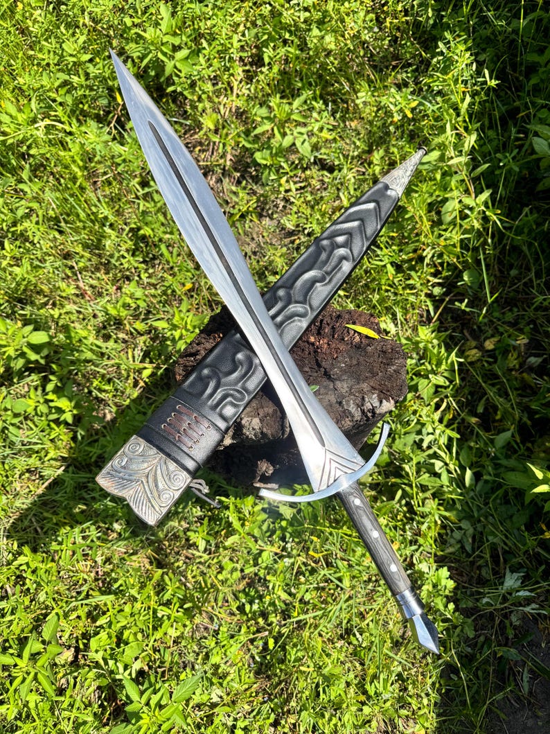 May include: A double-edged steel sword with a central fuller and a silver-toned crossguard. The handle is dark wood with metal rivets. It is accompanied by a black leather scabbard featuring embossed scrollwork, metal fittings, and leather lacing.