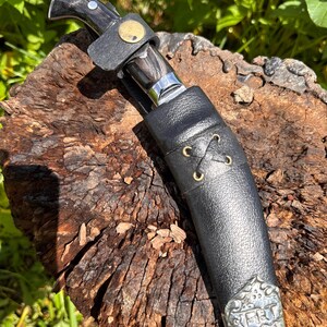 May include: A handcrafted knife with a dark wooden handle and a silver blade, secured in a black leather sheath. The sheath features decorative metal accents and a leather strap with a gold-colored button. The knife rests on a weathered wooden surface.