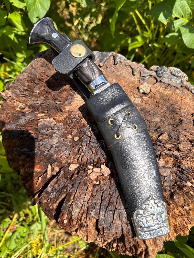 May include: A handcrafted knife with a dark wooden handle and a silver blade, secured in a black leather sheath. The sheath features decorative metal accents and a leather strap with a gold-colored button. The knife rests on a weathered wooden surface.