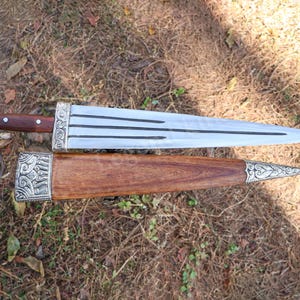 May include: A detailed image of a dagger with a wooden handle and sheath. The blade is silver with black lines, and the handle is dark brown. The sheath is also wooden, with silver decorative accents. The dagger is lying on a bed of dry leaves.