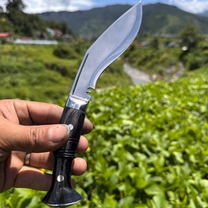May include: A kukri knife with a curved, polished steel blade and a black handle with silver accents. The knife is held against a backdrop of green foliage, mountains, and a blue sky.