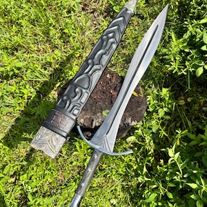 May include: A double-edged steel sword with a pointed tip, a central fuller, and a crossguard. The handle is dark with metallic accents. It is accompanied by a black leather scabbard featuring embossed patterns and a decorative silver-toned metal tip.