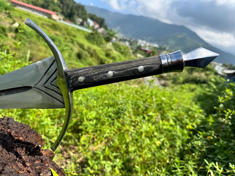 May include: A metal sword featuring a polished steel blade with engraved chevron details, a curved crossguard, and a dark wood handle secured with three silver rivets, finished with a pointed metal pommel at the end of the grip.