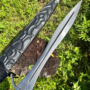 May include: A double-edged steel sword with a central fuller and a silver-toned crossguard. The blade is paired with a black leather scabbard featuring embossed chevron patterns and an intricate silver metal tip with engraved floral details.