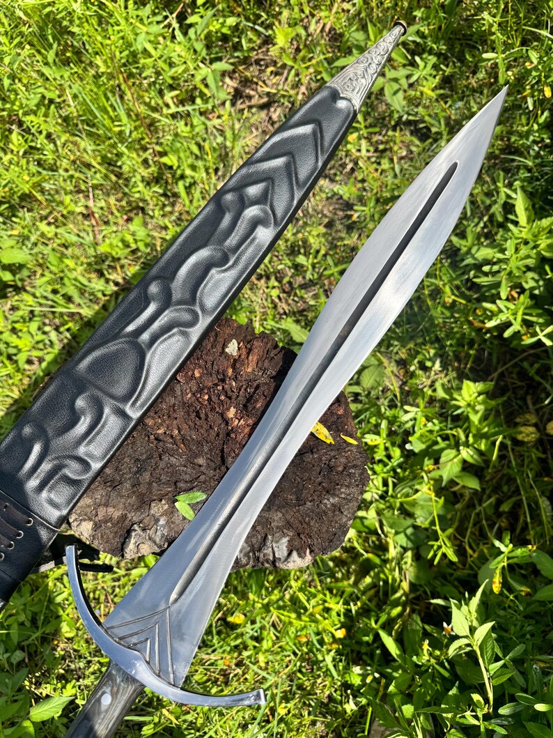 May include: A double-edged steel sword with a central fuller and a silver-toned crossguard. The blade is paired with a black leather scabbard featuring embossed chevron patterns and an intricate silver metal tip with engraved floral details.