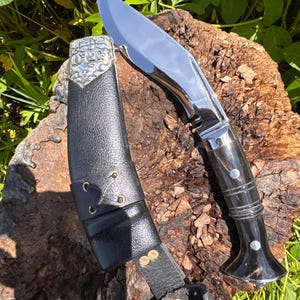 May include: A polished steel kukri knife with a curved blade and a black handle, resting on a wooden surface. The knife is accompanied by a black leather sheath with decorative metal accents and a belt loop. The blade reflects the surrounding environment.